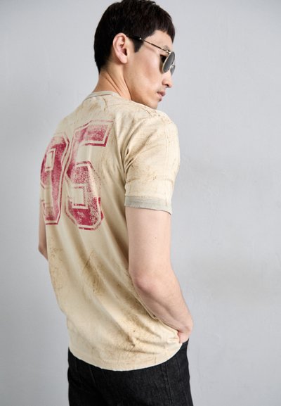 Cremfarbener T-Shirt mit verblasstem roten "95" Druck auf dem Rücken, leicht strukturierte Textur, kurze Ärmel, seitlich betrachtet.