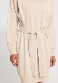 Robe en maille beige à manches longues avec ceinture en tissu assortie nouée à la taille, montrant des mains avec une bague en argent à la main gauche.