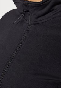 Chaqueta deportiva negra con cuello alto, que cuenta con una cremallera frontal y una tela suave y elástica. El diseño enfatiza una apariencia ajustada.