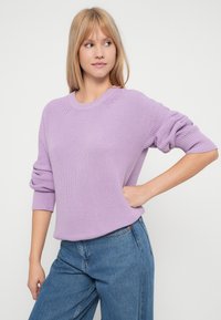 Mujer joven con cabello rubio lleva un suéter de punto lila y jeans azules, posando con una mano en la cadera frente a un fondo neutro.