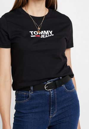 Kobieta w czarnej koszulce „Tommy Jeans”, nałożonych warstwowo złotych naszyjnikach, niebieskich dżinsach z czarnym paskiem z srebrną klamrą.