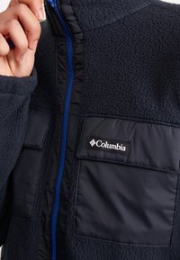 Granatowa bluza polarowa z niebieskim zamkiem błyskawicznym, posiadająca dwie kieszenie na piersi oraz logo Columbia. Miękka w dotyku, o przemyślanej konstrukcji.