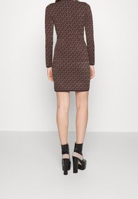 Robe à manches longues en tissu tricoté marron foncé présentant un motif logo sur l'ensemble. Le mannequin l'associe à des bottines à talons hauts noires et beiges.