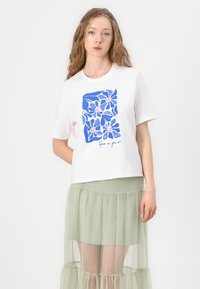 T-shirt blanc arborant un motif floral bleu et le texte "épanouis-toi tel que tu es". Associé à une jupe en tulle légère de couleur vert clair, superposée.