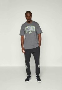 Grå bomulls t-skjorte med grønn og hvit "ADIDAS" grafikktrykk, sammen med svarte joggebukser med hvite side stripe og svarte sko.