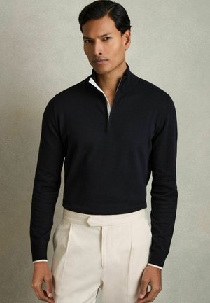 Schwarzer Zip-Kragen-Pullover aus weichem Stoff mit einem weißen Kragen-Detail. Kombiniert mit cremefarbenen, plissierten Hosen. Einfaches Design.