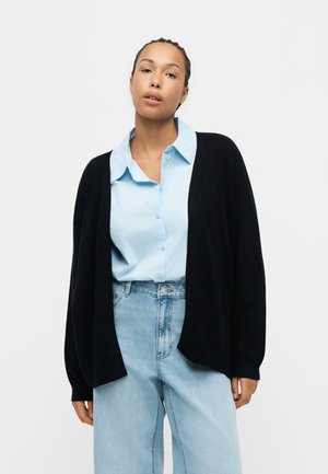 Kvinde står mod en ensfarvet baggrund iført en lyseblå skjorte med knapper, sort cardigan og lyseblå højtaljede jeans.