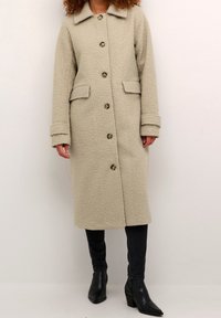 Beżowy płaszcz przypominający polar, z kołnierzem, długimi rękawami i zapięciem na guziki. Posiada dwie przednie kieszenie i teksturowaną tkaninę. Nosi się go na ciemnych dżinsach i butach.