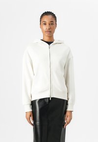 Felpa crema con zip, dotata di un grande cappuccio, tasche frontali e polsini a coste, abbinata a una gonna in pelle nera per creare un contrasto.