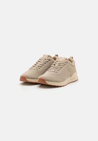 Zapatillas deportivas beige con parte superior de malla texturizada, punta redonda y suela de goma color coñac en contraste. Presentan cordones en tono a juego y una marca en la lengüeta.