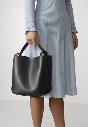Femme en robe bleu clair à manches longues tenant un grand sac cabas en cuir noir par ses poignées sur un fond uni.