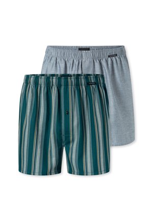 Hemdloser Mann mit lockigem dunklem Haar, der blaue und grüne vertikal gestreifte Boxershorts trägt, schaut nach unten und berührt die Rückseite seines Nackens.
