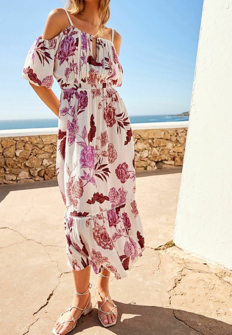 Femme portant une robe blanche à épaules dénudées à motifs floraux dans des tons roses et bordeaux, debout à l'extérieur près d'un mur en pierre avec l'océan en arrière-plan.