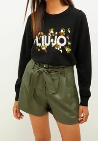 Suéter negro con el logotipo texturizado "LIU JO" en colores; combinado con pantalones cortos de cuero verde oliva de cintura alta con un cinturón atado y detalles de botones.