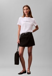 Fehér rövid ujjú póló "Calvin Klein Jeans" felirattal, fekete farmer szoknyával, fekete övvel és lapos fekete cipőkkel párosítva. Kézben táska.