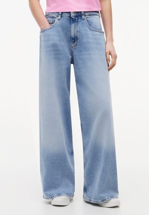 Jean bleu clair à jambes larges porté avec des baskets blanches et un haut rose, mains dans les poches avant, debout sur un fond blanc.