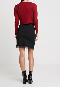 Woman wearing a cropped red sweater, black lace-trimmed skirt, and black ankle boots, standing facing away on a plain background.