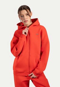 Sudadera con capucha y cremallera roja hecha de tela suave, con cremallera frontal, capucha grande y puños acanalados. Bolsillos laterales que ofrecen funcionalidad.