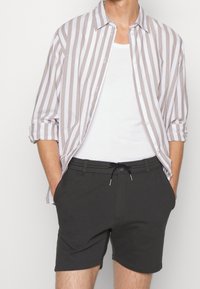 Chemise à rayures à manches longues de couleur beige et blanche, portée par-dessus un débardeur blanc. Associée à un short gris foncé avec une ceinture élastique et un cordon de serrage.