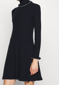 Mörkblå ribbad stickad klänning med hög krage, som har en utställd silhuett och vågiga kanter på ärmar och nederkant.