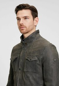 Schwarze Lederjacke mit hohem Kragen, zwei aufgesetzten Taschen auf der Brust, abgenutzter Textur und einem Wappenlogo am Ärmel.