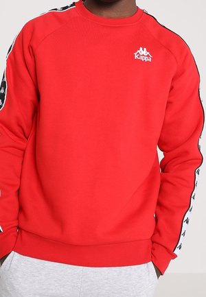 Czerwona bluza Kappa z czarnymi i białymi paskami z logo na rękawach, noszona przez osobę z rękami w kieszeniach jasnoszarych spodni.