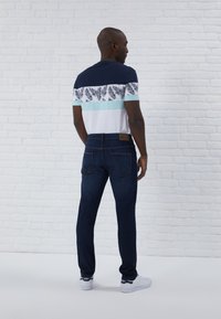 T-shirt pour homme en bleu marine avec une rayure blanche et verte à motifs de feuilles de palmier. Assorti à un jean en denim noir et des baskets blanches.