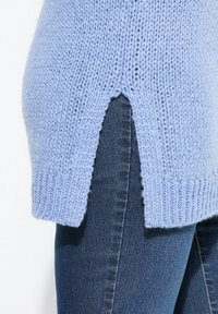 Světle modrý pletený svetr s rozděleným dolním lemem a žebrovanými manžetami, v kombinaci s tmavými džíny z denimu s hladkou texturou.