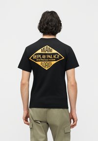 Άτομο με σκούρα σγουρά μαλλιά που φοράει μαύρο T-shirt με κίτρινο κείμενο "Miami Replay Palace 81st Marcoai Avenue Florida", συνδυασμένο με μπεζ παντελόνι τύπου cargo.