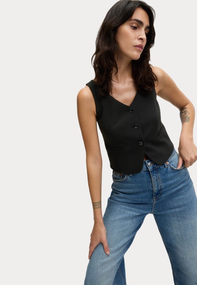 Gilet nero senza maniche con scollo a V e tre bottoni, abbinato a jeans blu a vita alta. Tessuto liscio con vestibilità su misura.
