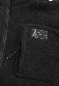 Chaqueta de forro polar negra con cremallera frontal y un bolsillo que presenta una etiqueta. La textura es suave y esponjosa, aumentando la calidez y la comodidad.