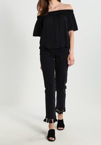 Haut noir à épaules dénudées avec un col froncé, associé à un jean court noir avec un ourlet effiloché et des accents de pompons. Talons ouverts noirs.