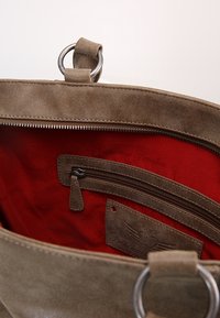 Brun mockaskinnväska med röd tygfoder, en dragkedjeficka och en etikett med präglad detaljering. Silverfärgade hårdvaru-detaljer ingår.