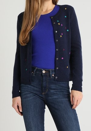 Woman wearing dark blue cardigan with small multicolored heart patterns over a bright blue ribbed top and dark blue jeans.