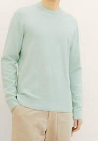 Person trägt einen hellblauen, strukturierten Langarmpullover und beige Hosen, mit einer Hand in der Tasche, vor einem schlichten Hintergrund.
