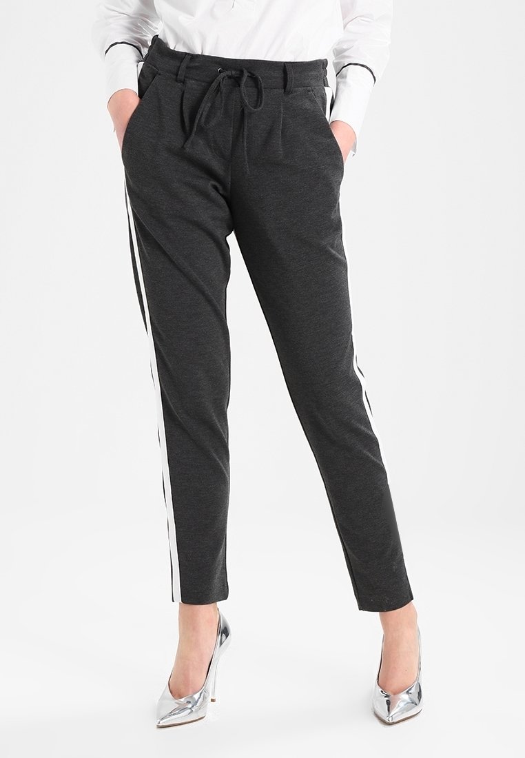 Pantalon gris foncé fuselé avec bandes blanches latérales, en tissu doux, avec taille à cordon et poches frontales. Talons hauts argentés visibles.