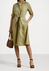 Abito in lino verde oliva con maniche corte, chiusura frontale a bottoni e vita legata. Abbinato a una borsa marrone e sandali con cinturini.