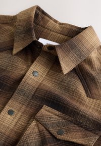 Chemise en flanelle à carreaux marron et foncé avec boutons-pression et poche poitrine avec rabat, présentée à plat.