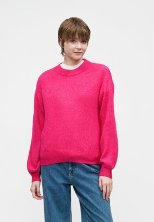 Jauns cilvēks ar īsiem brūniem matiem, ģērbies spilgti rozā džemperī un zilos džinsos, stāv uz vienkārša gaiša fona.