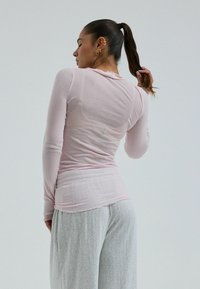 Žena s culíkem, která má na sobě světle růžové tričko s dlouhým rukávem a šedé texturované kalhoty, stojící v uvolněné póze před jednobarevným pozadím.