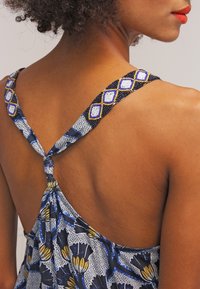 Gros plan sur le dos d'une femme portant une robe florale bleue avec des bretelles perlées nouées au centre, montrant ses cheveux bouclés et un profil avec du rouge à lèvres rouge.