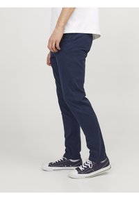 Vue de côté d'une personne portant un pantalon slim bleu marine, des chaussettes blanches, des baskets en toile bleu marine et un t-shirt blanc sur un fond uni.