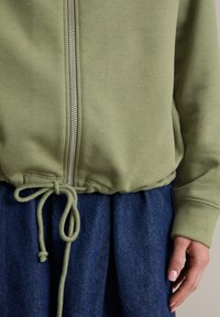 Olivegroene rits-hoodie met een trekkoord onderaan, geribbelde manchetten en een soepele textuur, gecombineerd met een donkerdenim rok.