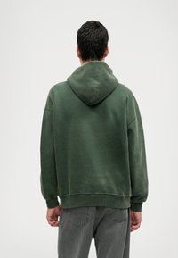 Tumši zaļš kapucēts džemperis ar brīvu piegriezumu, ribotām aprocēm un mīkstu tekstūru. Iekļauts kenguru kabata un noapaļots apakšmalas dizains.