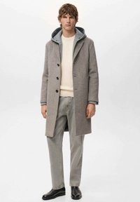Grijze wollen overjas met een grijze capuchon, voorzien van knopen aan de voorkant en zijzakken, gecombineerd met lichtgekleurde broek en zwarte laarzen.