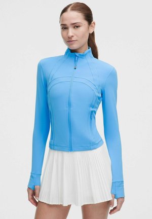 Femme portant une veste zippée bleu clair ajustée avec des trous pour les pouces et une jupe plissée blanche sur un fond uni.