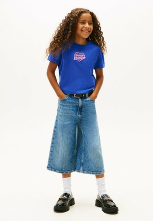 T-shirt en coton bleu avec le logo "Tommy Hilfiger", associé à des culottes en denim larges et des mocassins noirs. Chaussettes blanches visibles, tenue décontractée et relax.