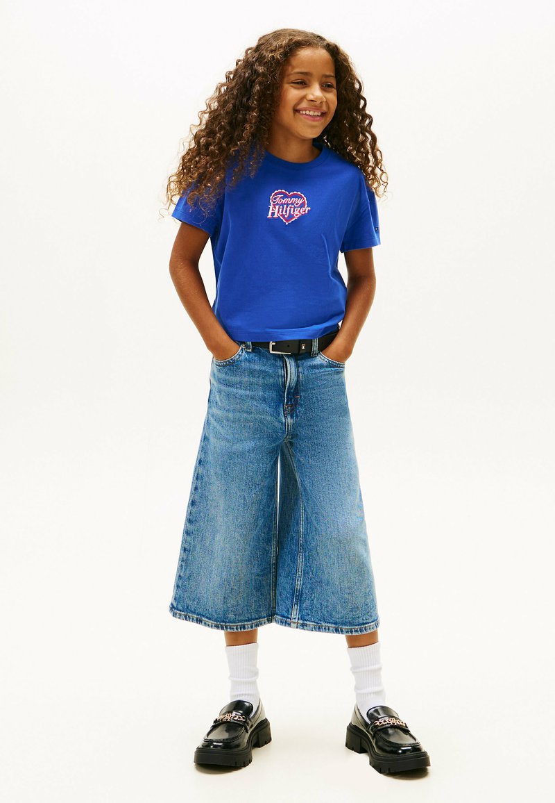 Camiseta de algodón azul con el logo de "Tommy Hilfiger", combinada con culottes de denim anchos y mocasines negros. Medias blancas visibles, atuendo casual y relajado.