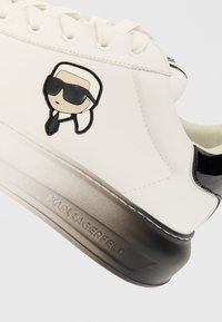 Bijela kožna tenisica s crnim akcentima, koja sadrži karikaturu loga s bočne strane, okrugle vezice i gradijentnu potplat s ugraviranim "KARL LAGERFELD".