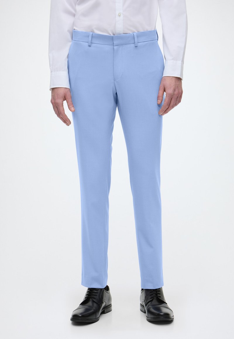 Pantaloni pe arcada de culoare albastru deschis, cu o textură netedă, două buzunare laterale și un talie cu bucle pentru curea. Purtați cu pantofi din piele negri.